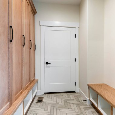 Beyaz yangın kapısı ve ringa kemiği desenli döşemesi olan kare bir Mudroom iç mekanı. Solunda ayakkabı deposu olan bir koltuk ve altında depo olan ahşap bir dolap var..