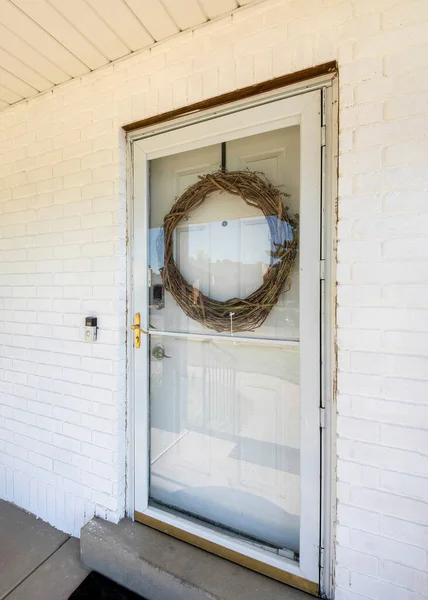 stock image Vertical Front exterior of a house with glass storm door over the white door with large leafless wreath. Home exterior with white bricks wall and ornaments outside near the bench.