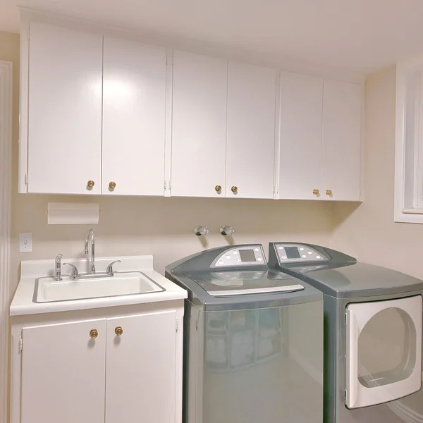 stock image Square Interior of a laundry room with an open white door and a view of the hallway. There are washer and dryer units beside the vanity sink under the wall cabinets beside the sliding windows on the left.