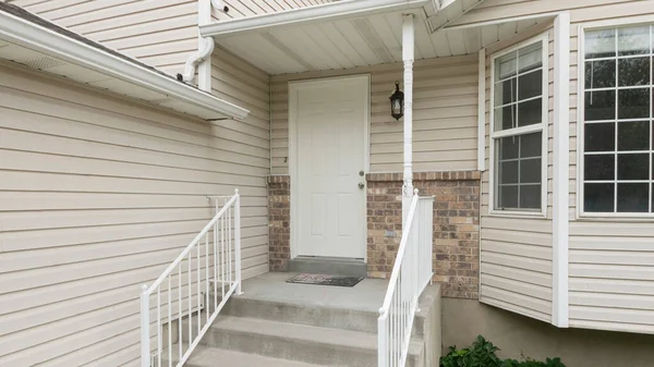 stock image Panorama Entrance of a house with white vinyl wood siding and bay windows. Front entrance of a house with railings at the doorsteps beside the bushes and grass on the left.