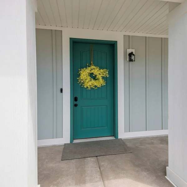 Square Front porch of a house with blue green front door and yellow wreath. Entrance exterior of a house with light gray board and batten sidings and two white column posts.