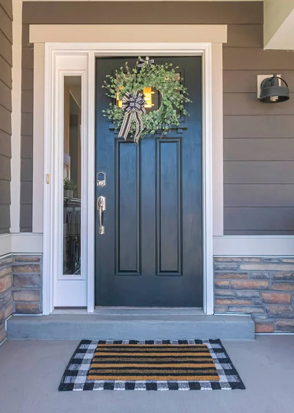 Vertical Black front door exterior with wreath and side panel. Entrance exterior with gray vinyl lap siding and woven armchair near the window on the right.