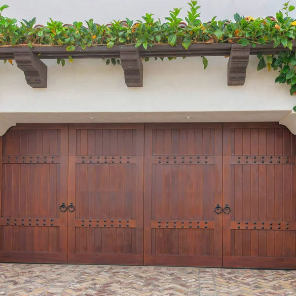 Square Dark wood side-hinged double garage doors at La Jolla, California. Exterior of a garage with crawling plants on the attached pergola and a driveway with bricks in herringbone pattern.