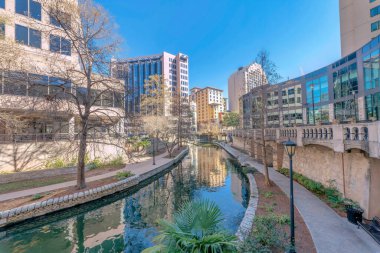 San Antonio River Walk Texas 'ta güneşli bir günde kanalın etrafı patikalarla çevrili. Dere boyunca uzanan binaların sakin su manzarası ve berrak mavi gökyüzü vardır..