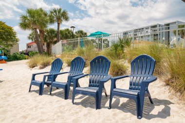 Destin, Florida. Plaj çimlerinin ve çitlerin yanındaki beyaz bir kumdaki dört şezlonglu sandalye. Arkaplanda bulutlu bir gökyüzünün altında binaların ve palmiye ağaçlarının manzarasına karşı boş koltuklar.
