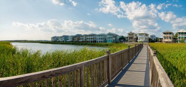 Destin, FL 'deki konutların yakınındaki gölde uzun otların üzerinde tahta korkuluklar olan tahta kaldırım. Gölün panoramik manzarası ve tahta kaldırım parlak gökyüzüne karşı yerleşim binalarına doğru gidiyor..