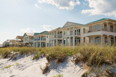 Destin, Florida- Sahildeki üç katlı evlerin önünde otlu beyaz kum tepeleri. Gökyüzüne karşı balkonları olan yazlık evler..