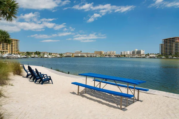 Dining table with seats and lounge chairs on a white sand shore at Destin, Florida bay. Chairs and dining table on a shore with lights and views of buildings and dock across the water.