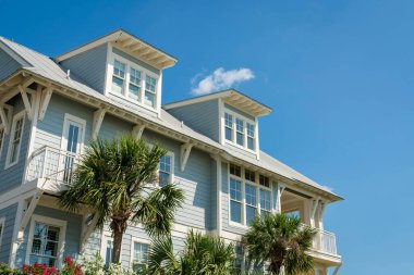 Destin, Florida 'da açık mavi ahşap kaplamalı ve beyaz çerçeveli bir evin alçak açılı görüntüsü. Evin duvarının yanında mavi gökyüzünün altında yatakhane çatıları olan palmiye ağaçları ve bitkiler var..