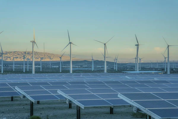 Desert wind farm near mountains with solar panels and wind turbines in California. View of solar panels and windmills during sunset and a background of mountain range sky at the back.