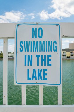 Destin, FL 'deki Gölde Yüzmek Yasaktır yazan beyaz korkuluklara işaret. Tuzlu suyla kaplı bir göl manzarasına karşı korkulukların üzerindeki tabelaya yakın çekim..
