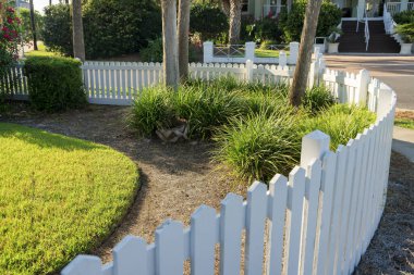 Destin, Florida. Kavisli köşeli beyaz çitlerle boyanmış. Çitin yanındaki çalıların ve ağaçların yanında solda budanmış çimenler var..