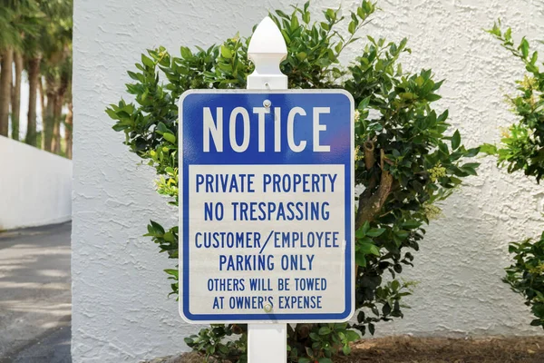 Destin, Florida- Sign with Notice Private Property No Trespassing near the bushes and wall. Sign on a post near the driveway on the left to a parking lot.