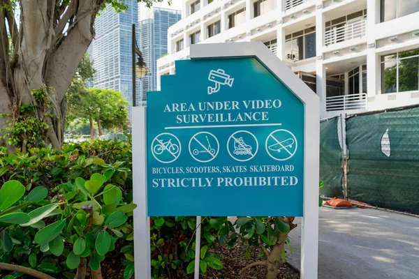 Area under video surveillance bicycles, scooters, skates, skateboard strictly prohibited sign- Miami. Signage near the plants and tree outside a building with chainlink fence with nets.