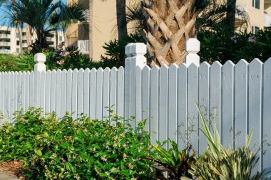 Destin, Florida... Boyalı beyaz çitlerin yanındaki bitkiler. Önünde yeşil bitkiler ve arka planında ağaçlar ve binalar olan açık hava çitleri..