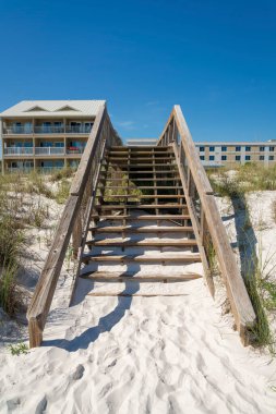 Destin, FL 'deki sahil evlerinin önündeki beyaz kum tepeciklerinin üzerindeki tahta kaldırımda merdivenler. Tahta merdivenler sahil evine çıkan bir kaldırım üzerinde gökyüzüne bakan binalar..