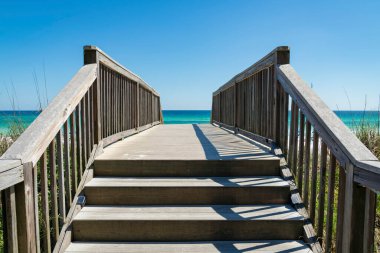 Mavi okyanus suyuna karşı bir yaya köprüsü ve Destin, Florida 'da açık bir gökyüzü. Merdivenler ve yaya köprüsü, aşağıdaki otların üzerinden plaja doğru giden tırabzanlar..