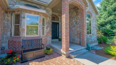 Panorama Whispy white clouds Exterior of a house with red bricks and stone veneer sidings. Front entrance of a house with arched entrance column posts and black front door near the windows on the right with bench. clipart
