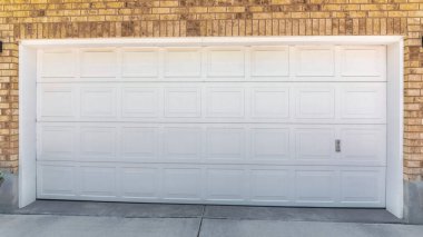 Beyaz bölmeli kapısı ve kahverengi tuğlaları olan bir garajın dışında. Evin garajında iki büyük saksıda bitkiler var. Kapının her iki tarafında da duvar lambaları var..