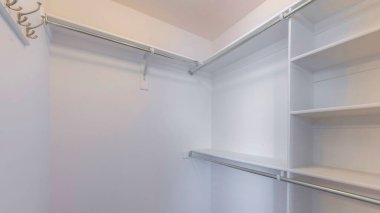 Kahverengi halı döşemeli Panorama küçük beyaz gömme dolap. Solunda metal çubuklar ve sağında kancalar olan bir raf ünitesi var..