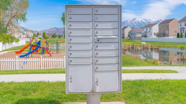 Panorama Whispy white clouds Cluster pedestal maibox near the playground and lake in a residential area at Utah valley. Community mailbox on a green lawn against the view of the playground, lake and houses at the back.