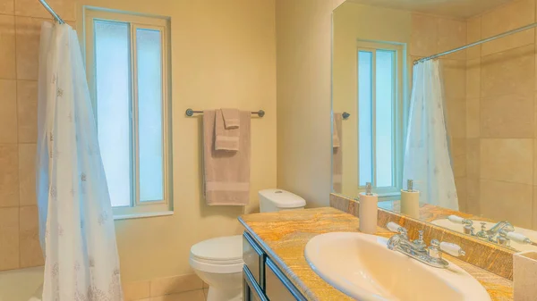 Panorama Bathroom interior with window and ambient lighting fixture. There is a vanity sink with granite top across beside the toilet with towels hanging on the side across the bathtub with shower curtain.