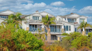 Panorama Beyaz Kabarık Bulutlu Çok katlı beyaz evler Carlsbad, California terasında cam parmaklıklar var. Önde yabani çalılar ve ağaçlar var ve arkada açık mavi gökyüzü var..