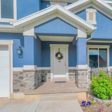 İki katlı bir bina. Bahçesi ve garajı var. Beyaz ön kapısı olan bir evin girişi sağdaki beyaz garaj kapısı ve soldaki bank..