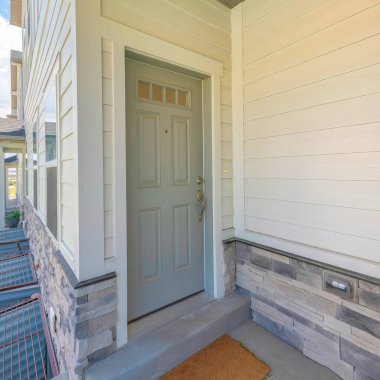 Camları ve paspası olan bir kasaba evinin gri kapısı. Beyaz vinil ahşap ve taş kaplamalı dış mekan girişi ve sağ taraftaki üç bodrum penceresinin manzarası..