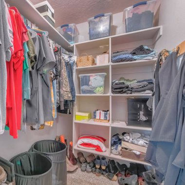 Square Small messy walk-in closet with a lot of shoes on the carpet floor. There are clothes on the hooks on the left and hanging clothes on the right near the cabinet with storage boxes and fabrics. clipart