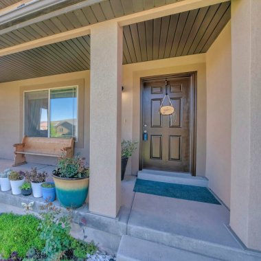 Önünde saksı bitkileri ve bahçe süprüntüleri olan bir evin ön cephesi. Pencerenin önündeki ahşap bankın yanında kahverengi ön kapılı ve tabelalı bir evin verandası..