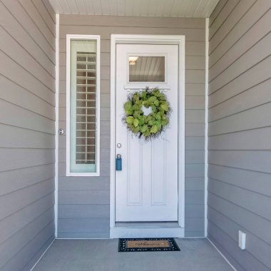 Gri ahşap kaplamalı ve ön kaplamalı taş kaplamalı bir evin kare girişi. Kilitli kutusu, çelengi, pencere paneli ve kenarı olan beyaz bir ön kapı var..