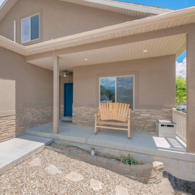 Açık kahverengi, iki katlı, garajı olan bir evin önündeki kare beyaz, kabarık bulutlar. Evin ön cephesinde ahşap bir bank ve verandada mavi bir ön kapı var solda garajın yanında sağda beyaz bir kapı var..