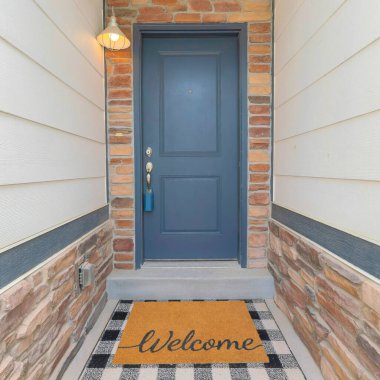 Duvarın ortasındaki kare küçük ev girişi bej ahşap vinil ve taş kaplamalı. Kilitli kutusu ve dekoratif paspası olan gri ön kapı..