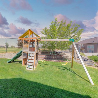 Gün batımında Kare Kabarık Bulutlar ahşap oyun alanı ekipmanları salıncak ve slaytlarla. Yeşil çimenlikte oyun parkı tel örgü çitlere ve arkadaki konut binalarına karşı..