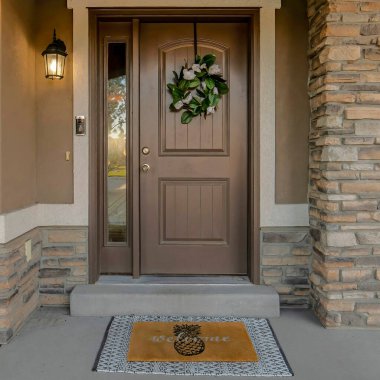 Kahverengi ve beyaz ana hatları olan bir evin ön kapısının önünde. Solunda çelenk ve yan paneli olan bir kapı ve sağında tabela olan boş bir tencere var..