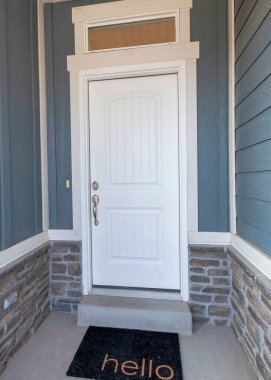 Ahşap duvar kaplamalı ve taş tuğlalı bir evin dış cephesi. Duvarın ortasında beyaz pencere ve siyah paspas olan bir duvar var..