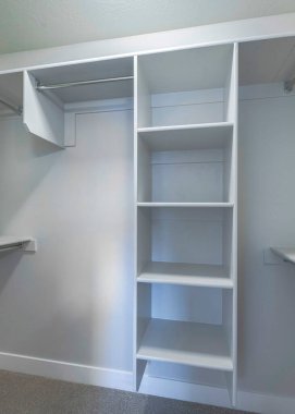 Dikey boş küçük penceresiz dolap ve açık gardırop. Duvarda metal çubuklar var beyaz ahşap raflar ve açık gri halılı döşemeler..