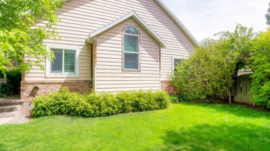 Panorama Front yard of a house with trees, plants and green lawn. Exterior of a house with entrance on the left and a wall with vinyl wood and bricks near the wooden gate on the right with plants above. clipart