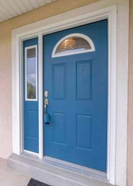 Dikey mavi ön kapı kilitli kutu, kemerli cam levhalar ve yan kapaklarla. Önünde çalılar olan beyaz çerçeveli, bej kaplamalı ve pencereli bir evin girişi.