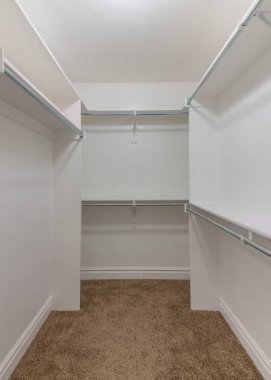 Dikey boş dolap raflarında metal çubuklar var. Dolabın içinde camsız bir yürüyüş. Duvarında beyaz raflar ve kahverengi halılar var..