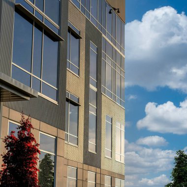 Gün batımında tuğlalarla, camlarla ve beton duvarlarla kaplı kare beyaz bulutlar bina cephesi. Binanın altında gökyüzü ufkuna karşı yeşil ve kırmızı ağaçlar var..