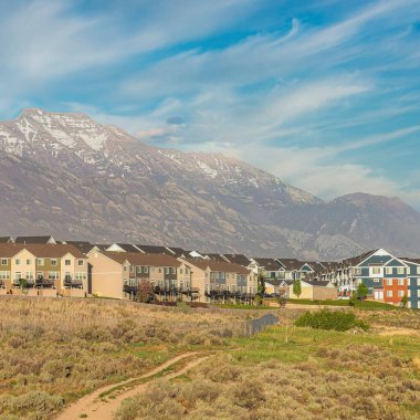 Kare Fısıldayan beyaz bulutlar geniş çalılar ve yerleşim alanının önünde toprak yolu olan çimenler. Dağ sırasına karşı renkli evler ve arka planda gökyüzü var..