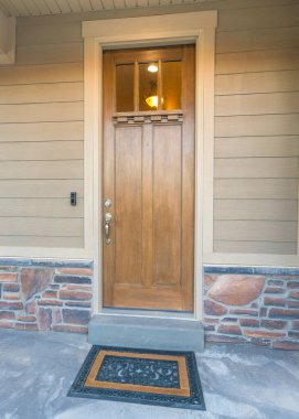 Cam camları ve şık paspası olan dikey ahşap ön kapı. Korkulukları ve bej vinil ahşap ve taş kaplamaları olan bir evin ön sundurması..