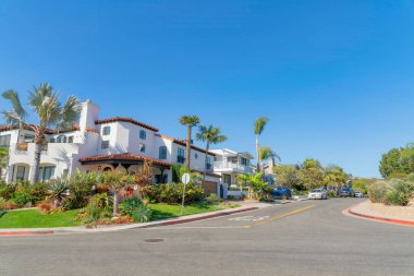 San Clemente, California 'da Akdeniz tarzı evleri olan bir yerleşim bölgesinde kavşak yolu. Solda büyük beyaz bir ev var. Palmiye ağaçları ve yol kenarında araçları olan çimenler var..