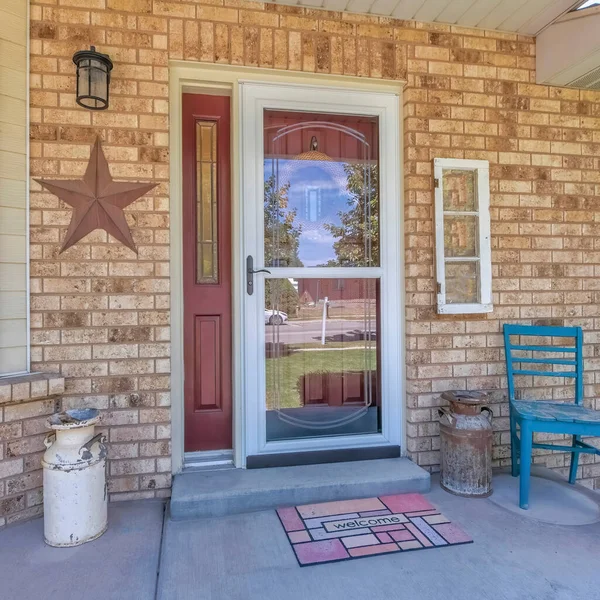 Square Home entrance with glass storm door over the burgundy door with side panel. Exterior of a house with bricks, windows and entrance with antique milk cans, chair and potted flowers.