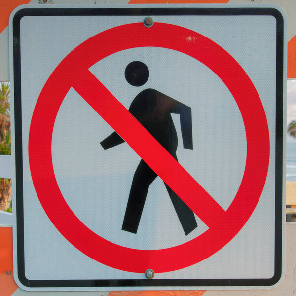 Square Puffy clouds at sunset No jaywalking sign on a white and orange barricade at Oceanside, California. Signage with a view of the beach waves, trees and fence at the back.