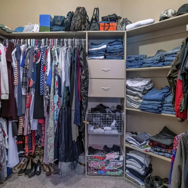Square Full organized walk in closet with open wardrobe and carpeted flooring. There are clothes hanging on the metal rods and a stacked of denims on the front wall with shoes at the bottom.