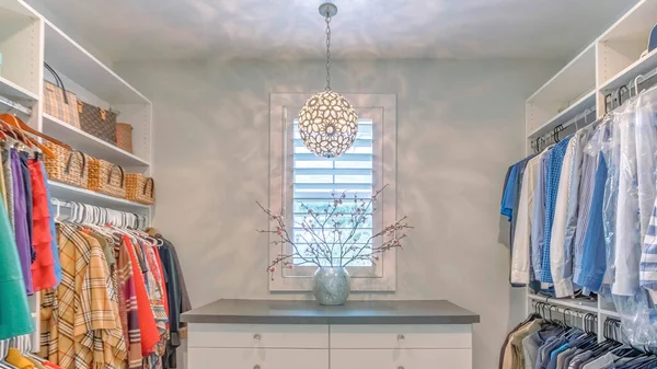 Panorama Large organized walk in closet with ball pendant lighting fixture. There are clothes hanging on both sides of the wall and a floor cabinet at the front with drawers and vase on top against the window.