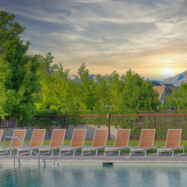 Square Whispy white clouds Lounge chairs on a public pool with wired fence at Daybreak, Utah. Community pool with steel handlebars on the ladders and a view of trees and houses against the mountain and sky background.